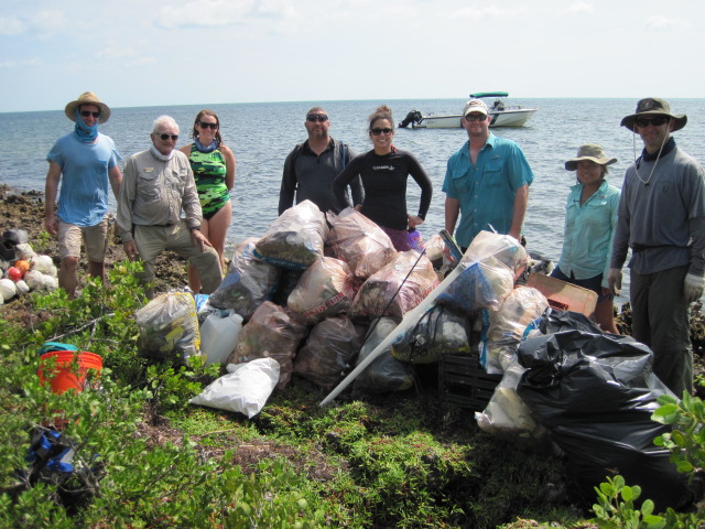 Eight people stand behind piles of garbage all stuffed in plastic garbage bags. A boat is floating on the water in the background.