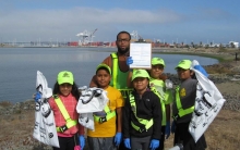 Kids and a chaperone on a beach with reflective gear and bags of debris and a city in the background.