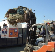 A person uses a forklift to life a large pile of fishing nets into a dumpster.