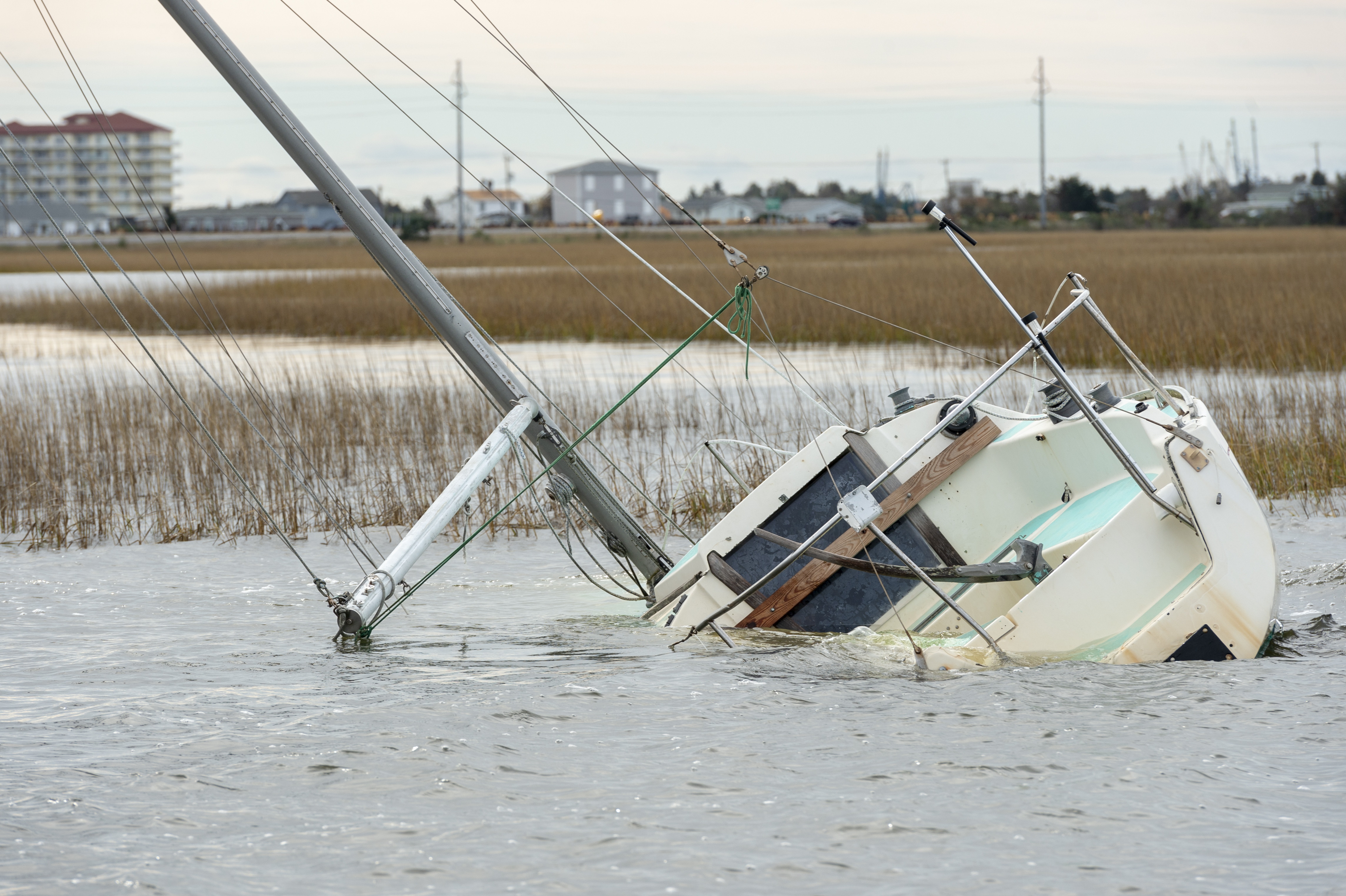 An abandoned and derelict vessel submerged in a marsh with buildings in the distance.