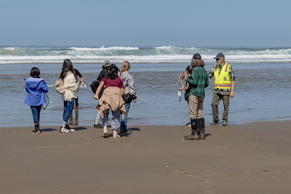 A group of young students holding a 5-meter rope stand on a sandy beach and look out into the waves.