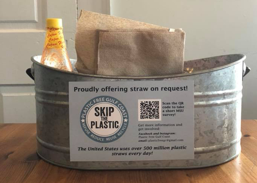 A metal tin sits on a wooden table and holds hot sauce and napkins. There is a sign on the tin that reads “Proudly offering straw on request! Skip the plastic.”