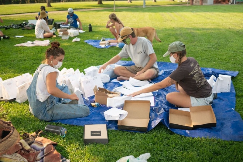 Volunteers wearing masks sitting on a tarp outside on the grass assembling school kits.