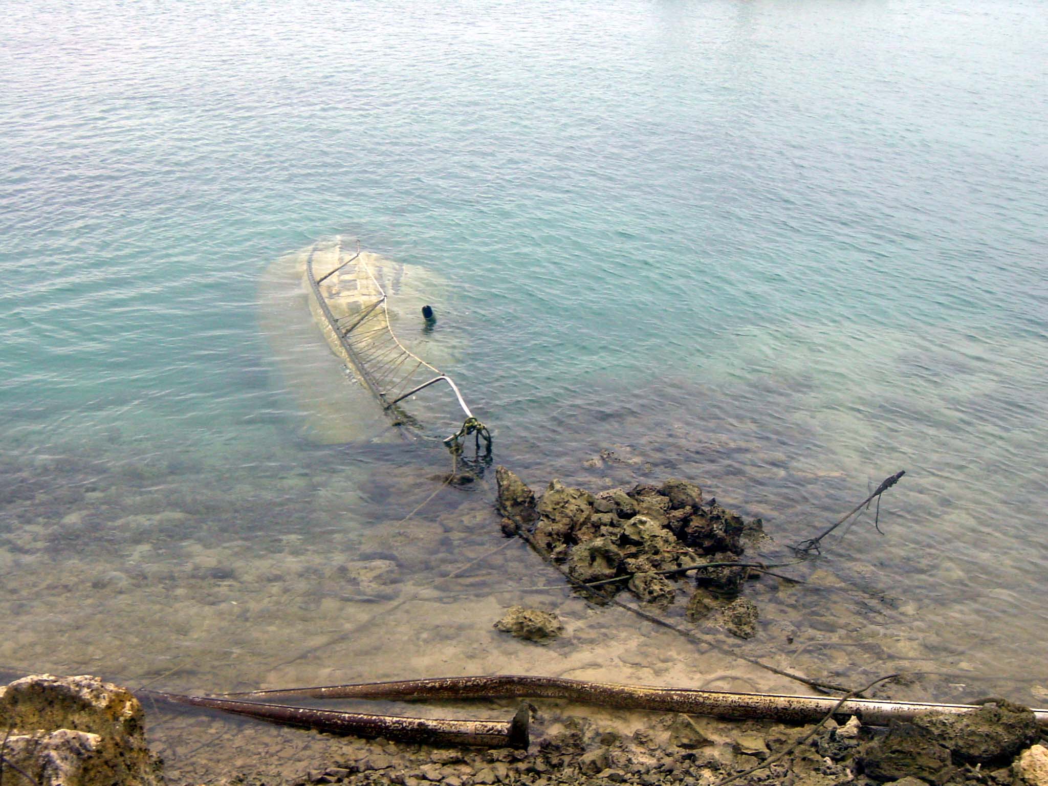 A sunken derelict vessel that is completely submerged in clear waters with the mast of the boat tangled in a rocky shoreline.