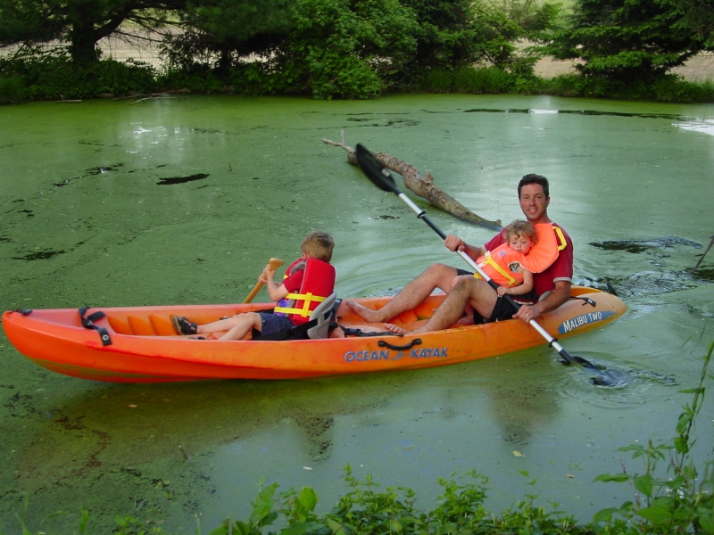 An adult and two children kayaking in a pond covered in algae.