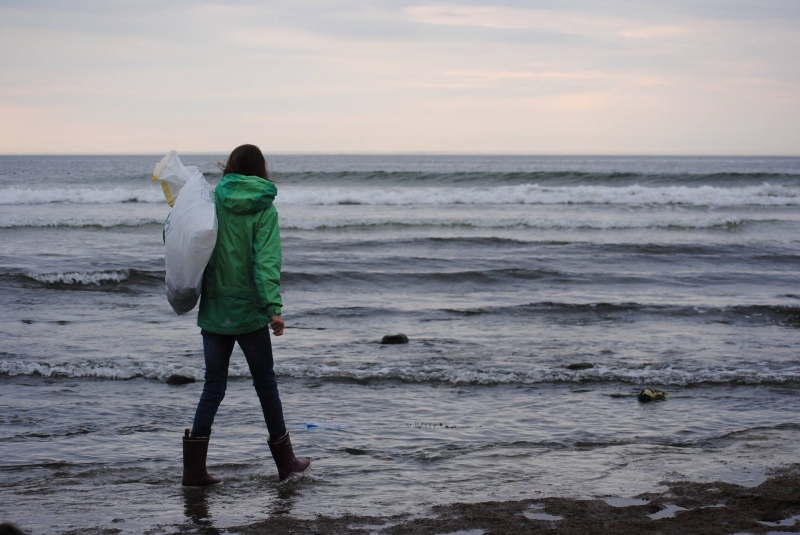 A woman walking along an ocean shore, carrying a bag of collected debris.