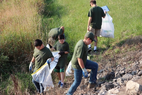 Five people hold plastic bags while they pick up trash on a shoreline.