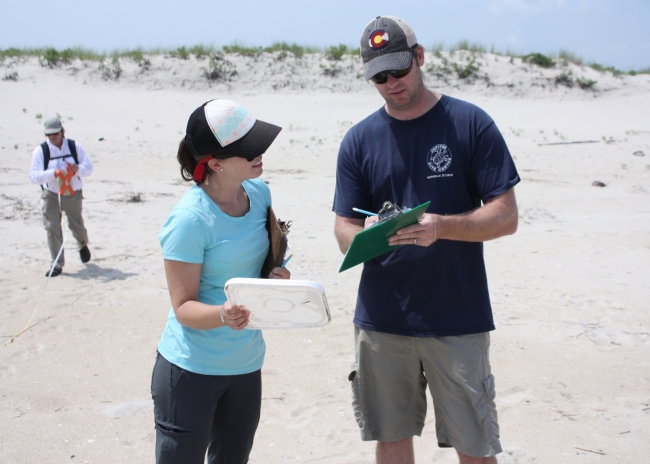 A woman holding a white plastic lid stands on a beach next to a man writing on a clipboard.