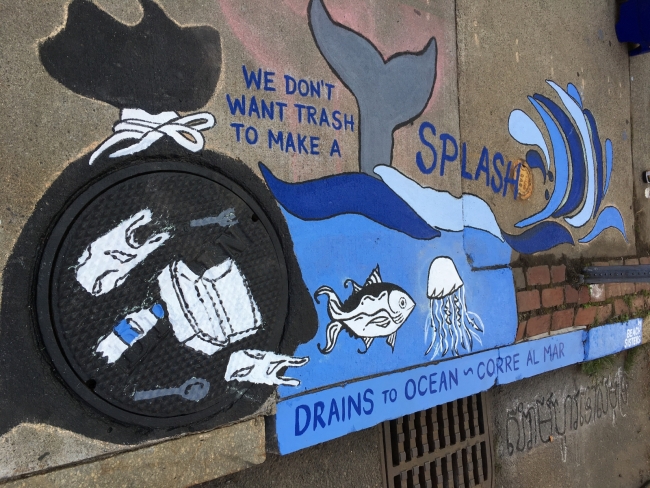 A painting over a sewer drain that says "We don't want trash to make a splash" and "Drains to Ocean" and shows an image of plastic trash.