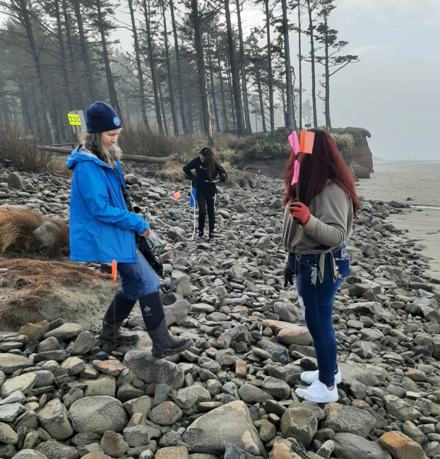 Two teen girls prepare to mark a transect with pink survey flags on a cobbled beach, guided by an older woman in a blue raincoat.