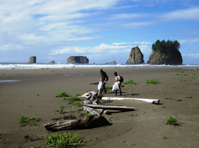 Two people walk along a long beach, carrying trash bags, with large rock mounts in the distance.