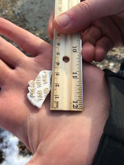 A student's hand holding a piece of plastic debris and measuring it with a ruler.