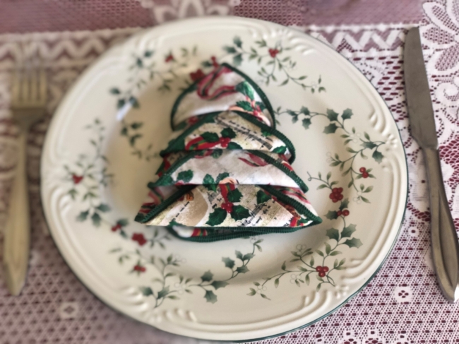 A cloth napkin is folded in the shape of a pine tree and sits on top of a glass plate. 