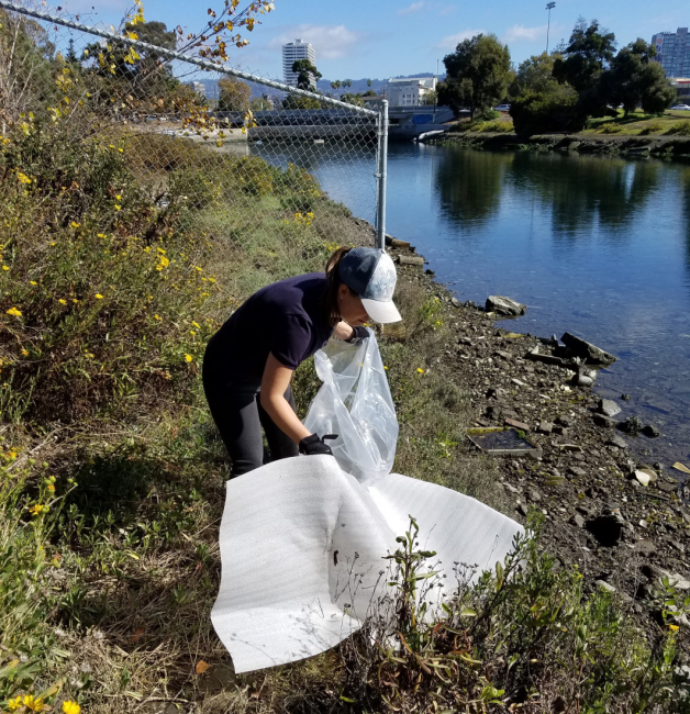 A volunteer picking up packing materials from next to a waterway with urban buildings in the background.