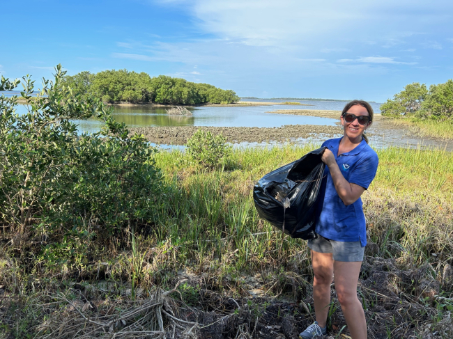 A volunteer holding a trash bag full of marine debris with a marsh ecosystem behind her.