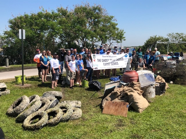 A group of people holds up a sign reading "The Ghost Trap Rodeo Event Series" behind a pile of tires and a pile of other debris.