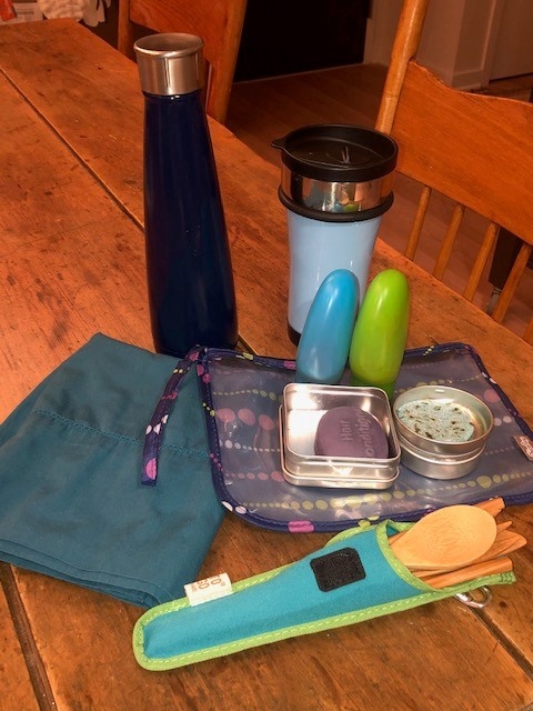 A cloth napkin, bamboo silverware, two reusable cups, and two shampoo bars sit on a table. 