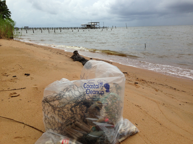 A large clear bag full of debris and labeled “Alabama Coastal Cleanup” is sitting on a sandy beach near the water’s edge.