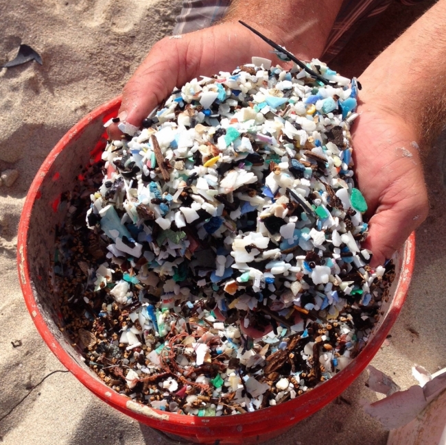 Two hands hold a pile of microplastics over the top of an orange bucket.