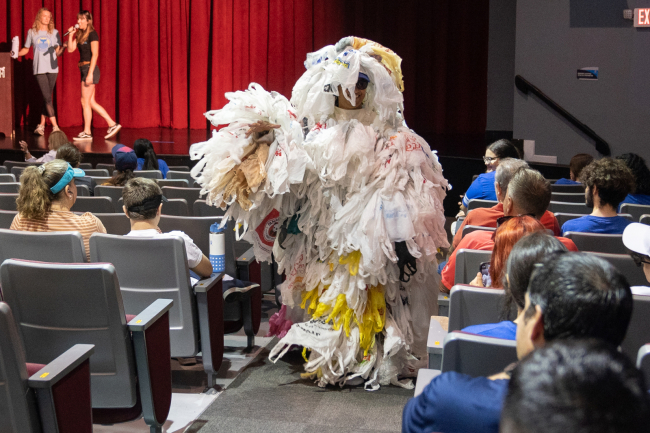 A person in a costume made of plastic bags walking through an auditorium audience.