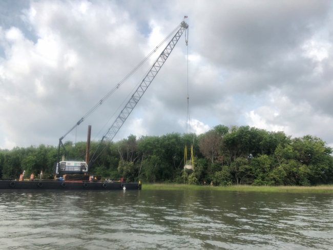 A Sailboat being lifted out of the marsh and maritime forest edge in the Charleston Harbor by a construction crane.