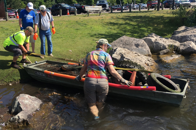 People pulling a boat ashore that is filled with marine debris collected along a lake shoreline.