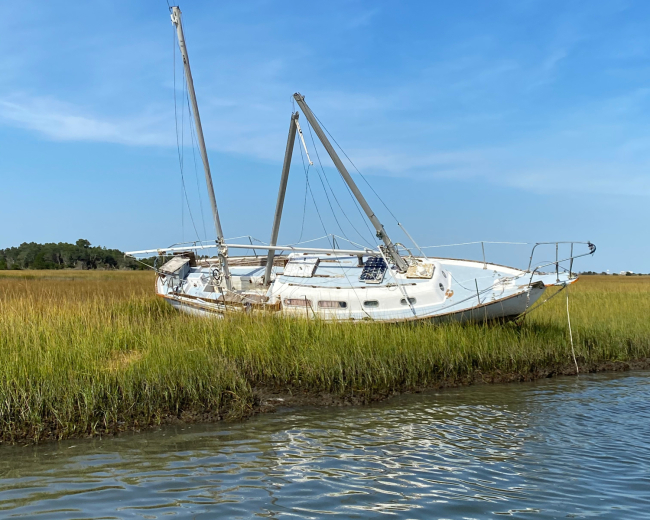 A derelict vessel on its side in a marsh along the water’s edge.