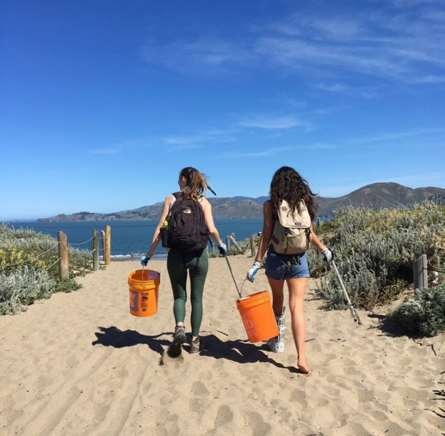 Two volunteers carry collected debris in buckets on a beach.