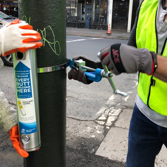 A metal cigarette butt disposal canister getting installed onto a pole along the street.