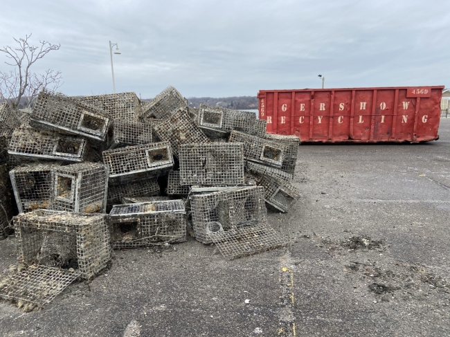 A pile of derelict lobster traps on a cement slab near a large red recycling dumpster.