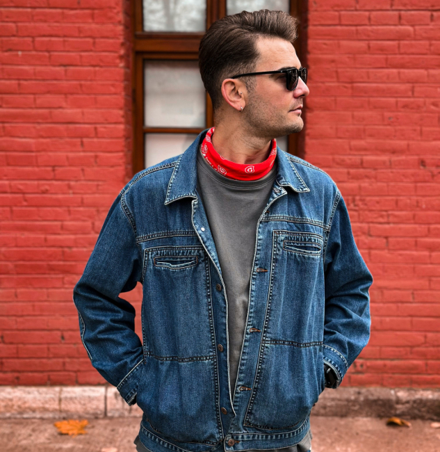 A man wearing a denim jacket and a red bandana against a brick building.