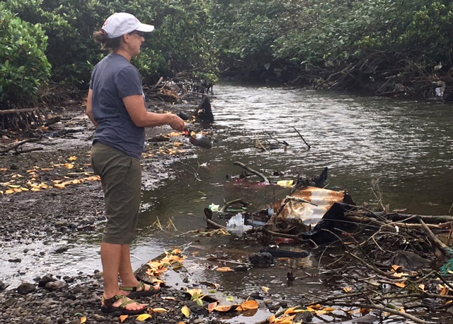 A person sampling sediment on the shore of a stream, near some trash tangled in natural debris.