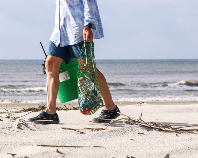 A volunteer collecting debris on the beach with a bucket and a bag made from old net material.