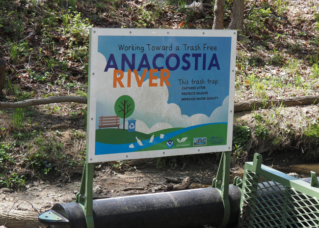 An informational sign titled "Working toward a trash free Anacostia River" on a litter trap installed on a stream.