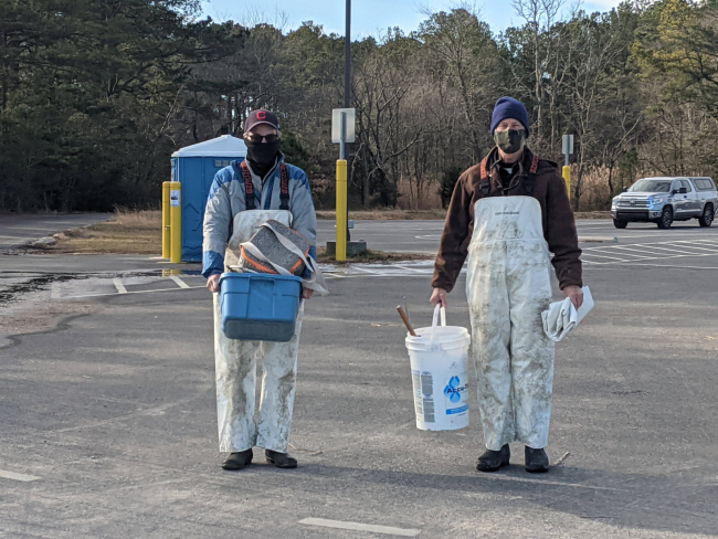 Two volunteers in dirty white coveralls carrying gear for a crab trap removal project.