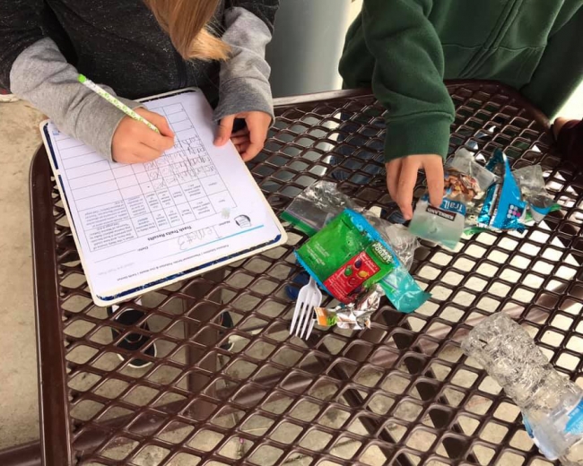 A student recording data on a clipboard next to food wrappers and trash on an outdoor table.