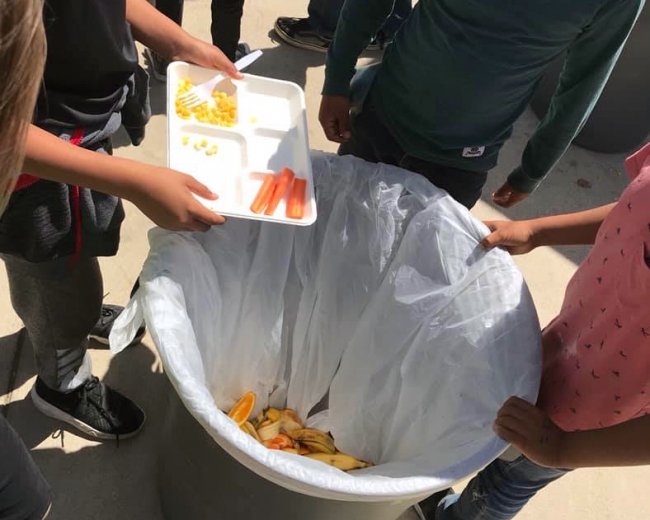 Students putting uneaten lunch food items into a large food waste bin.