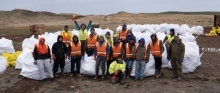 Seventeen people stand in a line for a picture with several white garbage bags full of marine debris.